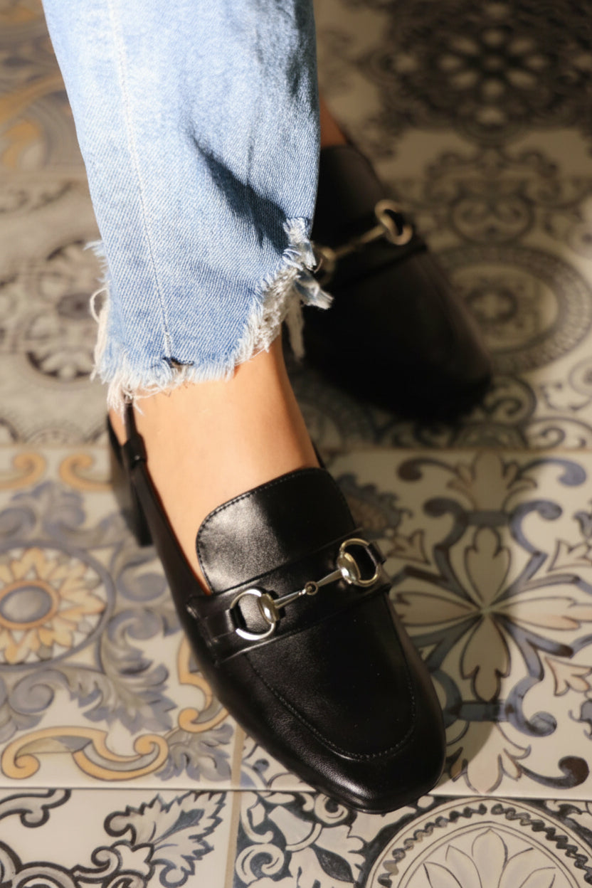 Black loafers worn with light blue jeans on a patterned floor.